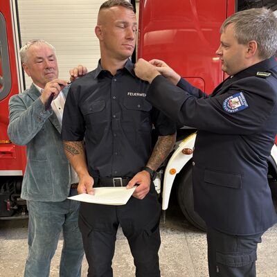 Bild vergr&ouml;&szlig;ern: Bürgermeister Thomas Beyer (l.) und Paul Wehry (r.), Leiter des Brandschutzamtes, befestigen neue Schulterstücke am Hemd von Hannes Zirnsack.
