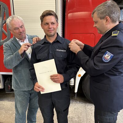 Bild vergr&ouml;&szlig;ern: Bürgermeister Thomas Beyer (l.) und Paul Wehry (r.), Leiter des Brandschutzamtes, befestigen neue Schulterstücke am Hemd von David Autenrieb.