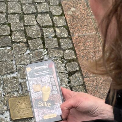 Bild vergr&ouml;&szlig;ern: Befinden sich Interessierte direkt am Stolperstein und scannen diesen per Smartphone-Kamera, werden auf dem Kamerabild Informationen in Textform zu der Person angezeigt, die an diesem Ort gelebt oder gewirkt hat. Anschließend können die Nutzerinnen und Nutzer Anteil nehmen, indem sie eine virtuelle Kerze am Stein platzieren. Die Kerze kann mit Namen und Botschaft personalisiert werden und ist auch für andere sichtbar. Die virtuelle Kerze brennt jeweils sieben Tage und kann danach erneut entzündet werden.